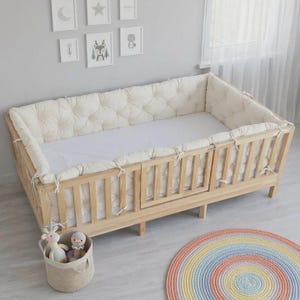 May include: A light-colored wooden crib featuring a tufted, off-white fabric bumper pad secured with ties. The crib has vertical slats and a white mattress. A round, multi-colored rainbow rug and a fabric basket containing two plush toys are nearby.