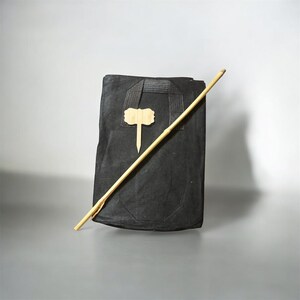 Puede incluir: Una bolsa tote negra con asa y una etiqueta de madera de color claro adjunta. Un palo de madera largo, delgado y de color claro descansa diagonalmente sobre la bolsa. La bolsa es rectangular y parece estar hecha de un material texturizado.