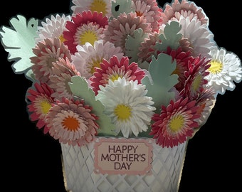 May include: A white basket filled with a bouquet of paper flowers. The flowers are in shades of pink, white, and green. The basket has a white label that says "Happy Mother's Day".