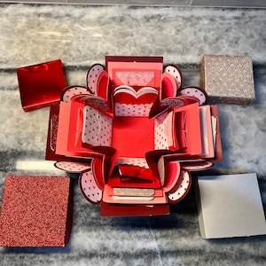 May include: A red and pink explosion box with a heart-shaped opening. The box is decorated with small white hearts and has a red glitter accent. The box is open and there are several smaller boxes inside.