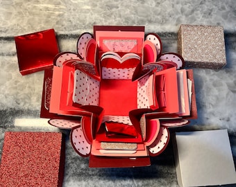 May include: A red and pink explosion box with a heart-shaped opening. The box is decorated with small white hearts and has a red glitter accent. The box is open and there are several smaller boxes inside.