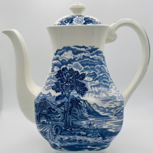 May include: A white ceramic coffee pot with a blue landscape design. The pot features a long spout, a curved handle, and a lid with a floral pattern. The landscape scene includes trees, a bridge, and figures near a body of water.