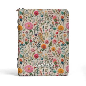 May include: A rectangular zippered planner cover featuring an off-white background with a colorful floral pattern of pink, yellow, orange, and blue wildflowers. The name Isabella is printed in a white script font near the bottom center of the cover.