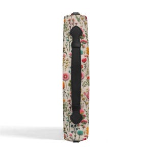 May include: A rectangular bag side panel featuring a cream-colored fabric with a colorful botanical print of various wildflowers in shades of pink, yellow, green, and blue. A vertical black leather handle is centered on the patterned fabric surface.