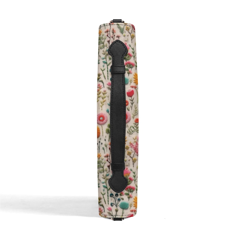 May include: A rectangular bag side panel featuring a cream-colored fabric with a colorful botanical print of various wildflowers in shades of pink, yellow, green, and blue. A vertical black leather handle is centered on the patterned fabric surface.