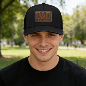 Puede incluir: Gorra de camionero negra con un parche rectangular marrón que dice "DAD EST.1994". La gorra tiene una parte trasera de malla y una visera curva. La persona de la imagen lleva una camiseta negra.