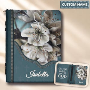 May include: A teal-colored Bible cover featuring a white floral print and the name Isabella in white script. The cover includes a vertical stitched seam and a subtle cross pattern. A smaller inset shows the back with the text Be Still AND KNOW THAT I am GOD.