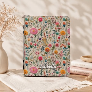 May include: A rectangular book cover featuring a cream-colored background with a dense, colorful pattern of embroidered-style wildflowers in shades of pink, orange, yellow, and blue. The name Isabella is printed in white script near the bottom.