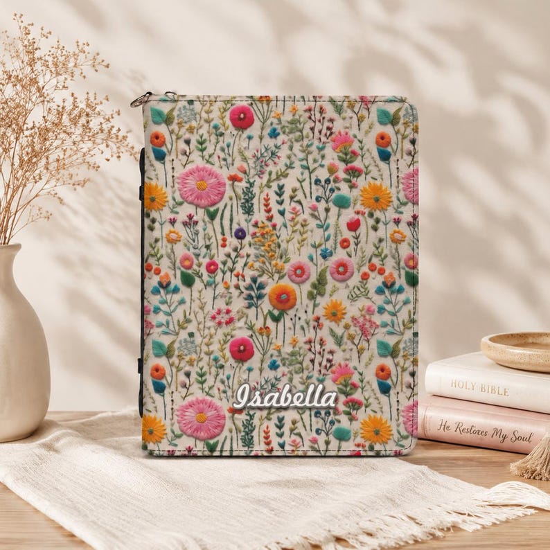 May include: A rectangular book cover featuring a cream-colored background with a dense, colorful pattern of embroidered-style wildflowers in shades of pink, orange, yellow, and blue. The name Isabella is printed in white script near the bottom.