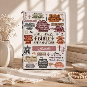 May include: A white Bible cover featuring a collage of brown and black religious text and symbols, including the phrase My Daily Bible Affirmations and the name Isabella in a script font, with a zipper closure and a black spine detail.