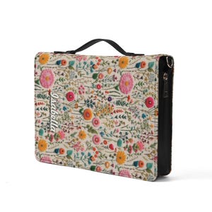 May include: A rectangular zippered case featuring a cream-colored fabric exterior with a vibrant, multi-colored floral embroidery pattern. The name Isabella is embroidered in white script on the left side. It includes a black top handle and zipper.