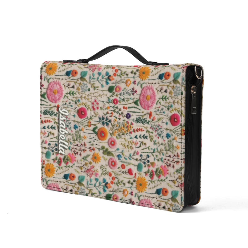 May include: A rectangular zippered case featuring a cream-colored fabric exterior with a vibrant, multi-colored floral embroidery pattern. The name Isabella is embroidered in white script on the left side. It includes a black top handle and zipper.