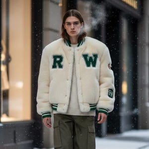 Op de afbeelding: Een crèmekleurige varsity jas met groene accenten en de letters "RW" op de borst. De jas wordt gecombineerd met een olijfgroene cargobroek en beige sneakers. De persoon loopt op een besneeuwde stoep.
