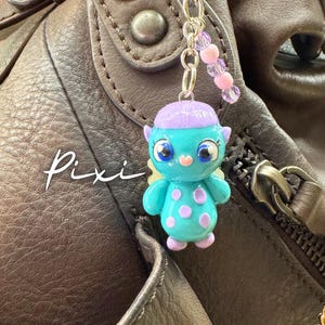 May include: A teal and purple Pixi charm with a beaded chain, hanging from a brown leather bag. The charm is a small, cartoon-like figure with large eyes and purple polka dots. The word "Pixi" is written in white script.