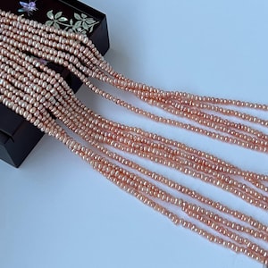 May include: Several strands of peach-toned freshwater pearls are displayed, emanating from a dark wooden box. The small pearls are uniform in size, forming a graceful, flowing arrangement. The box features a decorative floral pattern on its lid.
