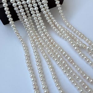 May include: Several strands of white pearls cascade from a dark wooden box. The pearls are round and have a lustrous sheen. The image is well-lit, highlighting the smooth texture and color of the pearls. The pearls are likely used for jewelry.