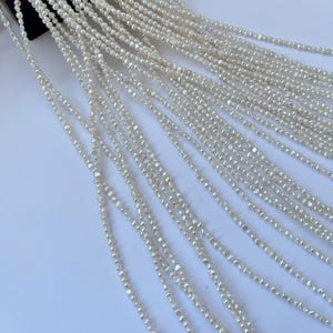 Op de afbeelding: Close-up van meerdere strengen kleine, gebroken witte, facetgeslepen kralen. De kralen zijn aan elkaar geregen, waardoor een watervaleffect ontstaat. De kralen lijken gemaakt van een reflecterend materiaal, dat het licht opvangt en een subtiele glans creëert. De achtergrond is een effen wit oppervlak.