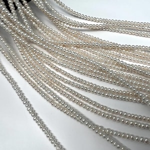 May include: Multiple long strands of small, white, round, lustrous pearls arranged in parallel lines. Each strand consists of numerous tiny, uniform beads strung together, creating a continuous, flowing texture of white, reflective, spherical objects.