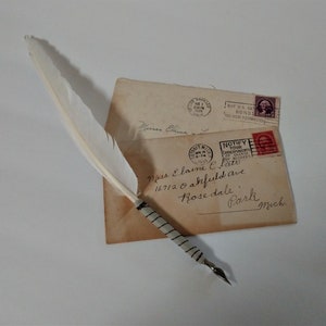 May include: A white feather quill pen with a black and white striped handle resting on top of two envelopes. The envelopes are addressed to Miss Elaine C. Clats, 16712 Oakfield Ave, Rosedale Park, Mich. The envelopes have stamps from the United States Postal Service.