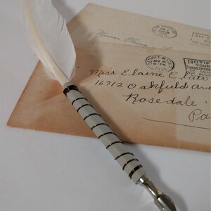 May include: A white feather quill pen with a silver and black metal nib and a silver and black wire wrapped handle. The pen is resting on a vintage envelope with handwritten text and several postage stamps.