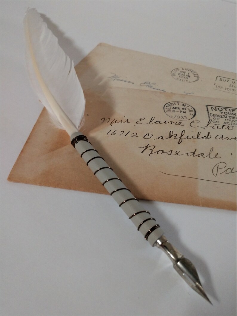 May include: A white feather quill pen with a silver and black metal nib and a silver and black wire wrapped handle. The pen is resting on a vintage envelope with handwritten text and several postage stamps.