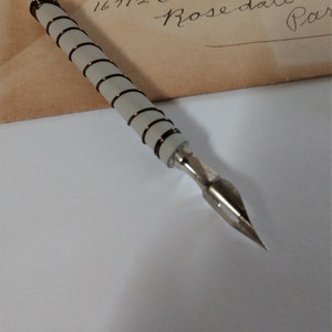 May include: A white feather quill pen with a gray and brown wrapped handle and a silver nib. The pen is resting on a vintage envelope with handwritten text that reads "Miss Elaine E. lator 16712 Oakfield ave. Rosedale, Par"