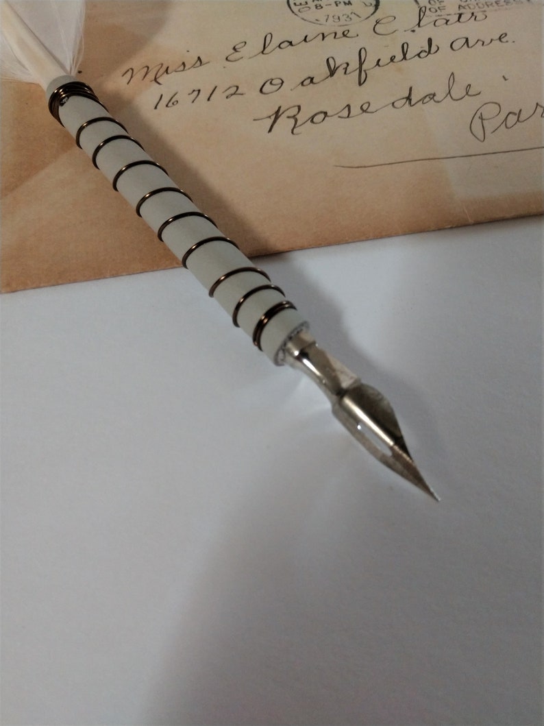May include: A white feather quill pen with a gray and brown wrapped handle and a silver nib. The pen is resting on a vintage envelope with handwritten text that reads "Miss Elaine E. lator 16712 Oakfield ave. Rosedale, Par"