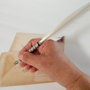 May include: A hand holding a white feather quill pen with a black and silver band over a vintage envelope with handwritten text.