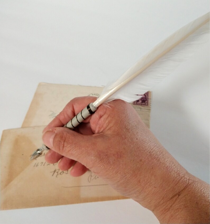 May include: A hand holding a white feather quill pen with a black and silver band over a vintage envelope with handwritten text.