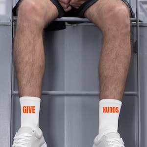 Könnte beinhalten: Weiße Socken mit dem Wort "GIVE" in Orange auf einer Socke und "KUDOS" auf der anderen. Die Socken werden mit weißen Turnschuhen getragen. Das Bild zeigt eine Person, die auf einer Metallkonstruktion sitzt.