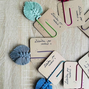 Peut inclure: Plusieurs marque-pages faits à la main en macramé, de couleurs variées comme le turquoise, le bleu et le bordeaux. Chaque marque-page est orné d'une feuille en macramé, d'un trombone coloré et d'une petite carte avec du texte manuscrit.