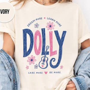 Könnte beinhalten: Ein elfenbeinfarbenes Kurzarm-T-Shirt mit einem Grafikdruck, der den Text DOLLY in großen blauen und rosa Buchstaben zeigt. Das Design enthält eine kleine akustische Gitarre, Blumen, Sterne sowie die Phrasen DREAM MORE, LEARN MORE, CARE MORE, BE MORE.