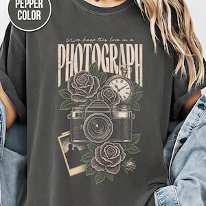 以下が含まれることがあります： ペッパーカラーのTシャツ。ヴィンテージスタイルの写真デザイン。デザインにはカメラ、バラ、懐中時計、「We keep this love in a PHOTOGRAPH」の文字が含まれています。