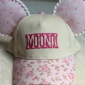 May include: A beige baseball cap featuring the word MINI embroidered in pink block letters on the front. The brim has a pink floral pattern, and two large, round, pink patterned ears with white pom-pom trim are attached to the top of the cap.