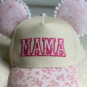 May include: A beige baseball cap featuring the word MAMA embroidered in pink block letters on the front. The brim has a pink floral pattern, and two round, light pink ears with white pom-pom trim are attached to the top of the cap.