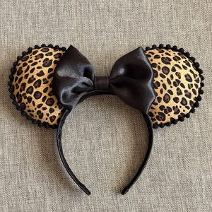 May include: A black headband with two leopard print ears and a large black bow. The ears have a brown and black spotted pattern with a black scalloped trim. The bow is made of a shiny black material.