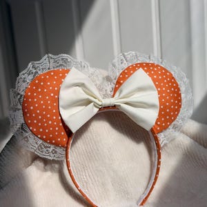 May include: A headband featuring two round orange ears with white polka dots, each bordered by white lace trim. A large, solid off-white bow is centered between the ears. The headband itself is covered in matching orange fabric with white polka dots.