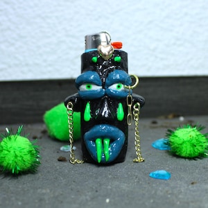 Könnte beinhalten: Ein schwarzes Feuerzeug mit einem geformten Gesichtsdesign. Das Gesicht hat blaue Lippen, grüne Zähne und grünäugige, blau geschminkte Augen. Goldketten hängen von den Seiten. Ein kleines goldenes Herz und ein roter Akzent schmücken die Oberseite. Ein einzigartiges, künstlerisches Accessoire.