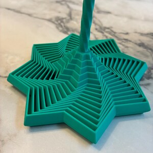 Pode incluir: Uma base para copos em forma de estrela turquesa com um design geométrico. A base para copos tem uma construção em camadas, com cada camada ligeiramente deslocada para criar um efeito escalonado. Uma haste torcida turquesa eleva-se do centro.