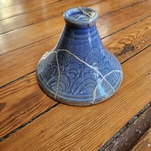 May include: A blue ceramic bowl with a wide base and a narrow neck, featuring a wave pattern. The bowl is broken, with visible cracks filled with a gold-colored material. The bowl sits on a wooden surface.