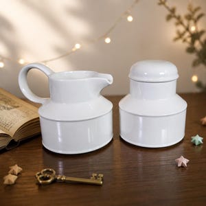 May include: A white ceramic creamer pitcher with a curved handle and a spout, paired with a matching white lidded sugar bowl. Both pieces feature a smooth, glossy finish and a tiered, cylindrical shape, standing side by side on a dark surface.