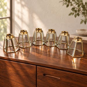 May include: A set of six glass and gold metal geometric lamp shades. Each shade features a hexagonal shape with clear glass panels framed by a metallic gold finish, topped with a circular gold rim, designed for use as a decorative light fixture.