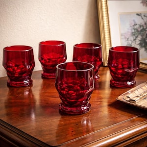 May include: A collection of six ruby red drinking glasses with a textured, faceted design. The glasses are arranged on a polished wooden surface, with a framed picture in the background. The glasses are empty and ready to be filled.
