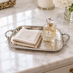 May include: A rectangular silver tray with ornate handles and a beaded rim holds a folded beige linen napkin with an embroidered leaf motif and a square glass perfume bottle labeled Eau de Parfum with a faceted crystal stopper.
