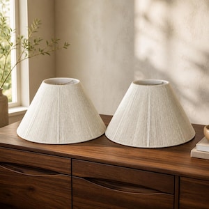 May include: A pair of matching off-white lamp shades with a conical shape and a vertical ribbed texture. Each shade features a wide circular base and a narrower top opening, constructed with fine, tightly wound thread detailing throughout the exterior.