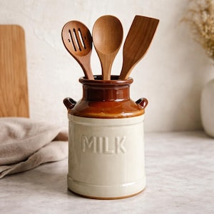 May include: A ceramic milk jug-shaped utensil holder with three wooden kitchen tools. The jug is cream-colored with a brown top and the word "MILK" embossed on the front. The wooden tools include a slotted spoon, a solid spoon, and a spatula.