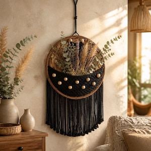 May include: A circular wall hanging featuring a woven rattan frame and a black macrame pocket. The pocket is adorned with light-colored wooden beads and holds dried eucalyptus and pampas grass. Long black fringe hangs from the bottom.