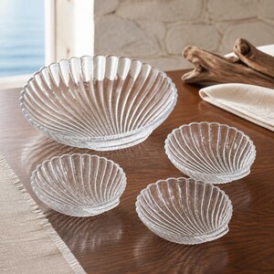 May include: A set of four clear glass dishes shaped like seashells, featuring a textured surface with radiating ridges. The collection includes one large bowl and three smaller bowls, all crafted from transparent glass with a scalloped edge design.