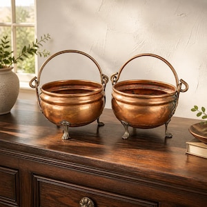 May include: Two matching copper pots with rounded bodies, each featuring a curved metal handle attached to the sides and three small, ornate feet at the base. The pots have a polished metallic finish with visible warm orange and brown tones.