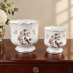 May include: Two white porcelain planters with scalloped rims and gold-painted edges. Each vessel features a colorful botanical design with a bird perched on a flowering branch, accented by gold star-shaped motifs on a glossy white surface.